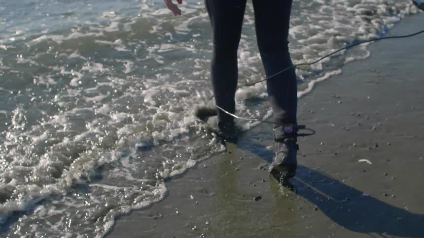 A surfer attached to his surfboard with a leash is wading out into the surf