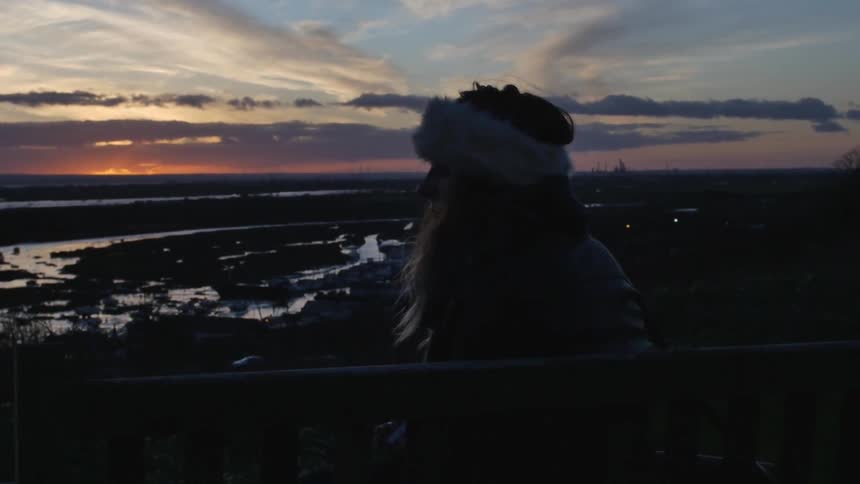 Woman standing and watching ahead as the sun sets in a cloudy sky
