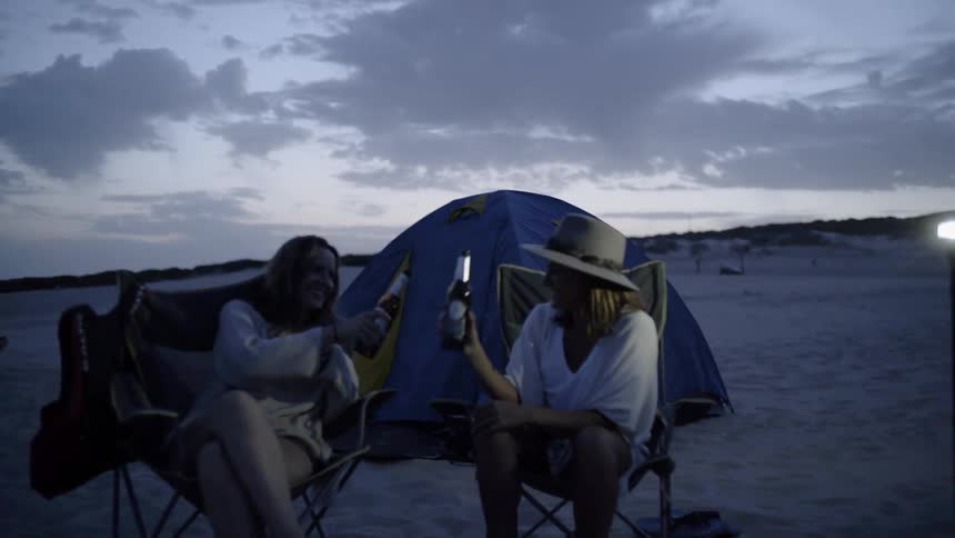 Two women share a toast as they sit at a campsite in the desert at dusk