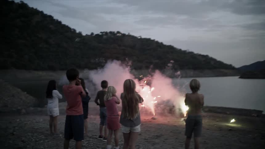 A group of children are standing on a beach watching firecrackers explode