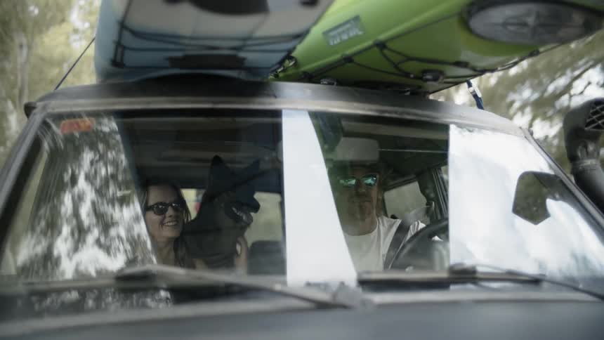 A man, woman and dog are driving in a car with kayaks on top