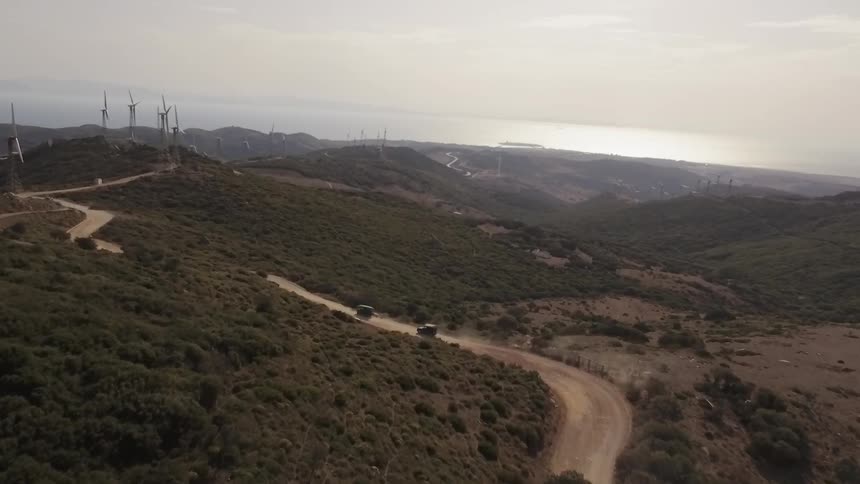 Vehicles travel on a dirt road toward a hilltop with wind turbines during the day