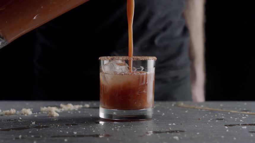 Someone is pouring a red liquid into a clear liquid inside of a rocks glass filled with ice
