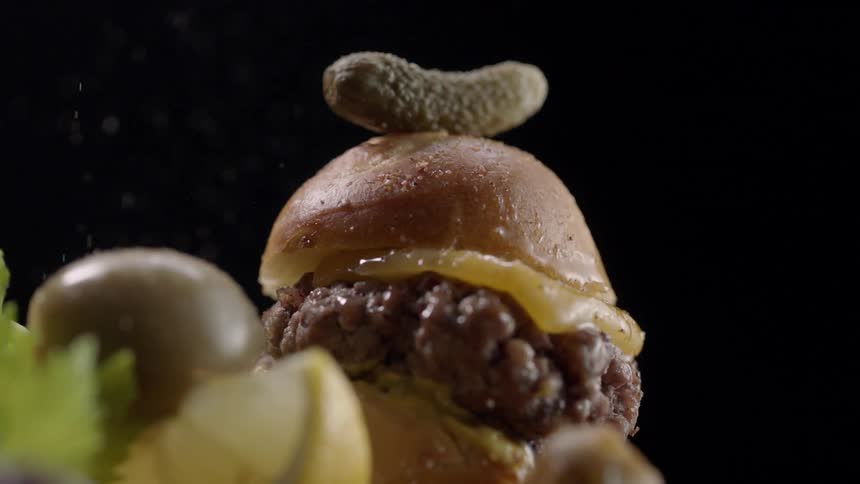 A burger that has a pickle on top of the bun is getting salt sprinkled on it