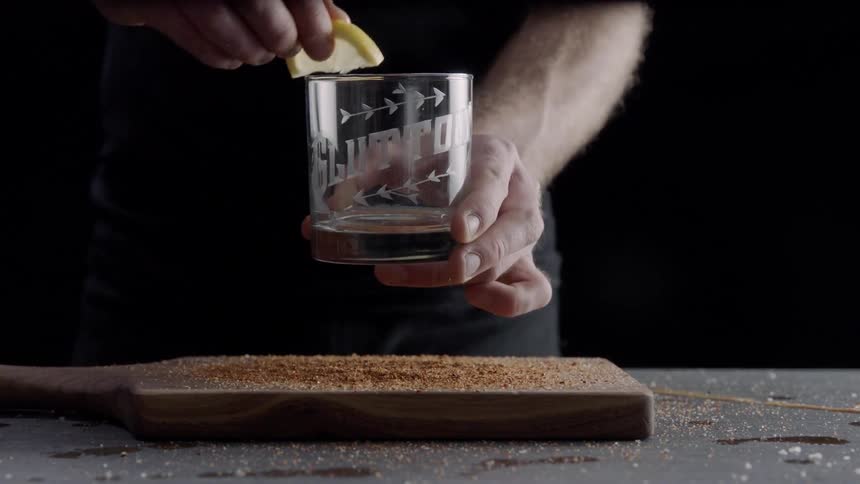 Someone is rubbing a lemon around the top of a rocks glass