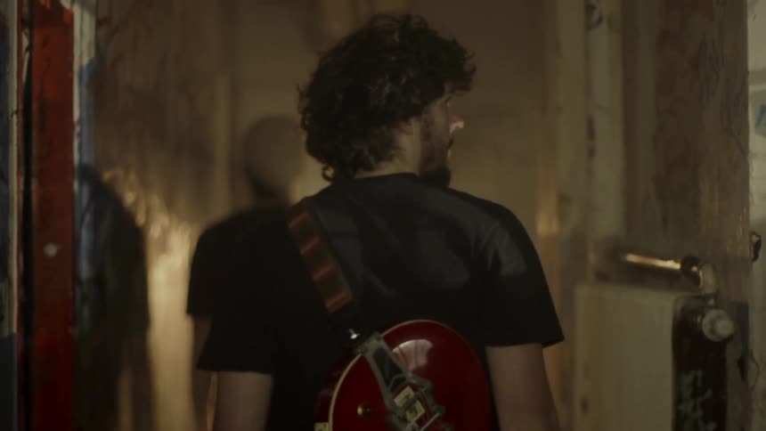 Man with a guitar slung over his back walks down a hallway