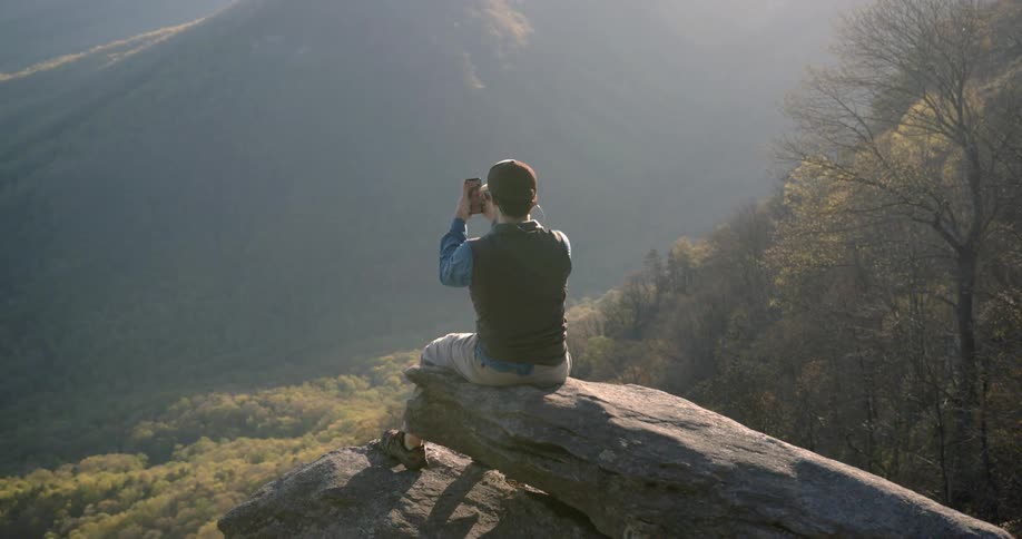 A man is sitting on the edge of a cliff and taking a picture with his phone