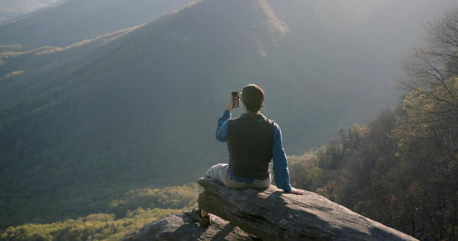 A man is sitting and taking pictures of a mountain view from a precipice