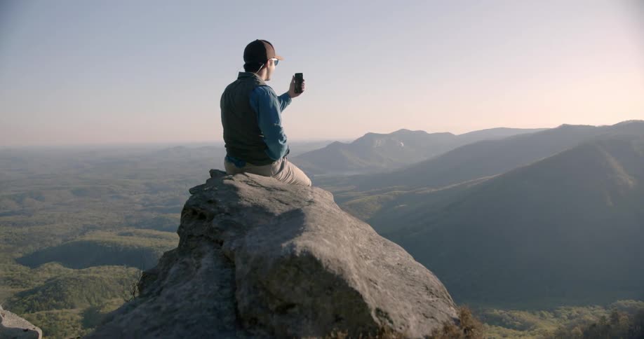 Man sitting on top of mountain recording video on cell phone of surroundings