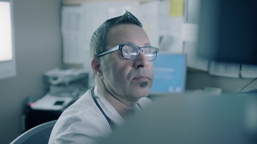 A male doctor is looking at computer monitors at a desk in an office