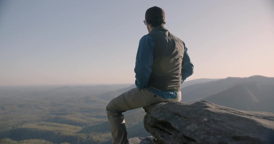 A man is sitting on the ledge of a cliff looking down into a valley