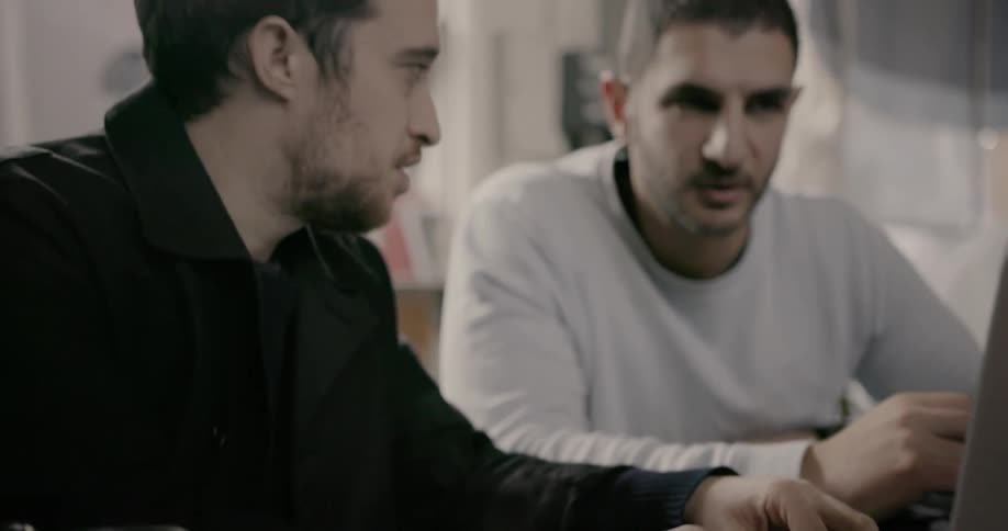 Two men are talking and working on a laptop as they sit at a coffee counter during the day