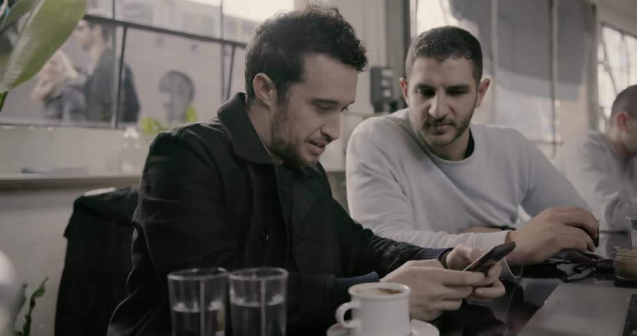 Men sit in a cafe and talk as one types into his smartphone
