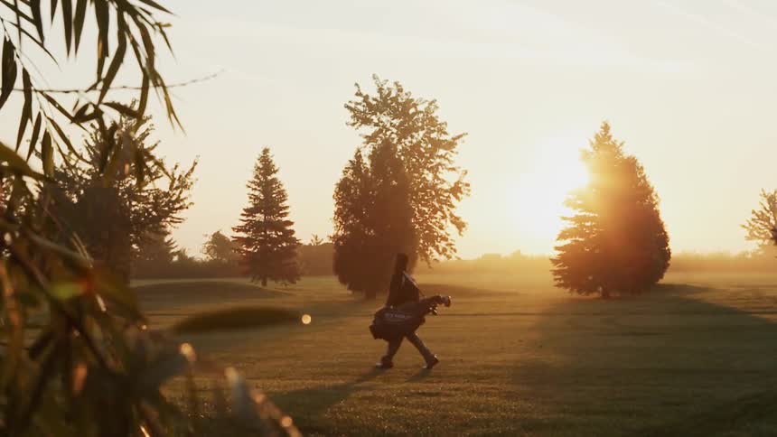 Sun is rising as a person with golf clubs walks past the green