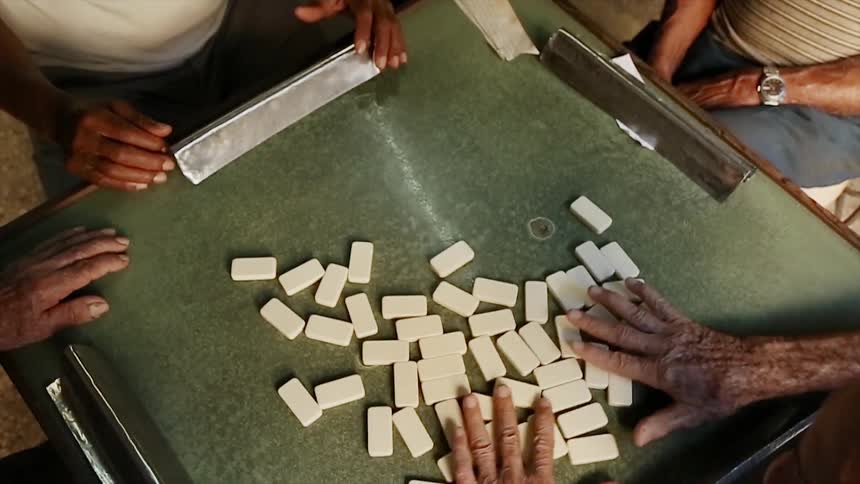 Men sit at a table while one of them shuffles dominoes