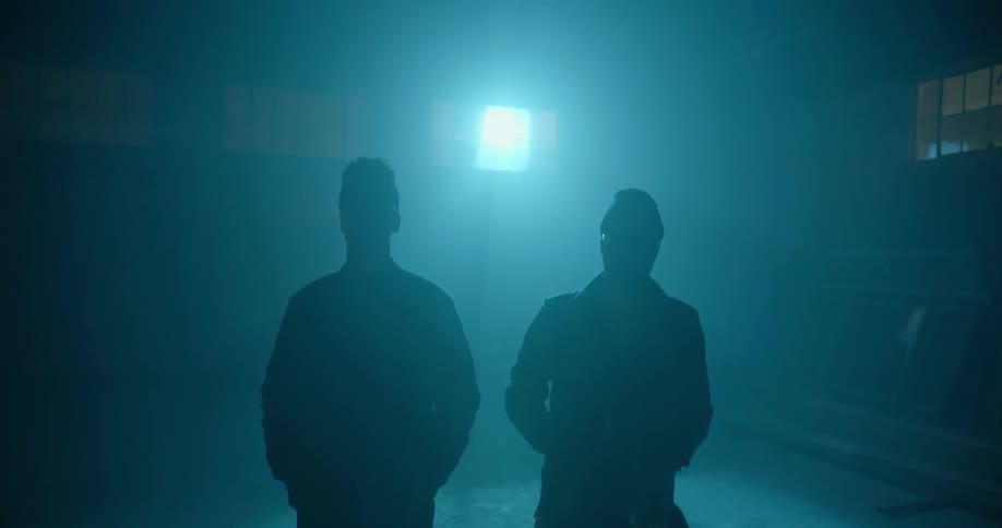 Two people stand still in a dark room with a single light