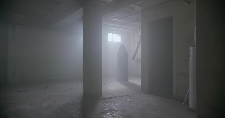 A cloaked figure stands in an empty basement room with a window