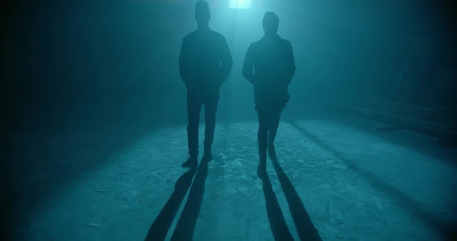 Two men are walking in a dimly lit warehouse during the day