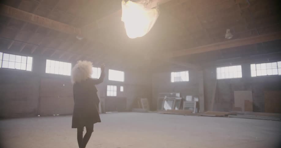 Woman walks across empty warehouse floor and reaches toward a spotlight