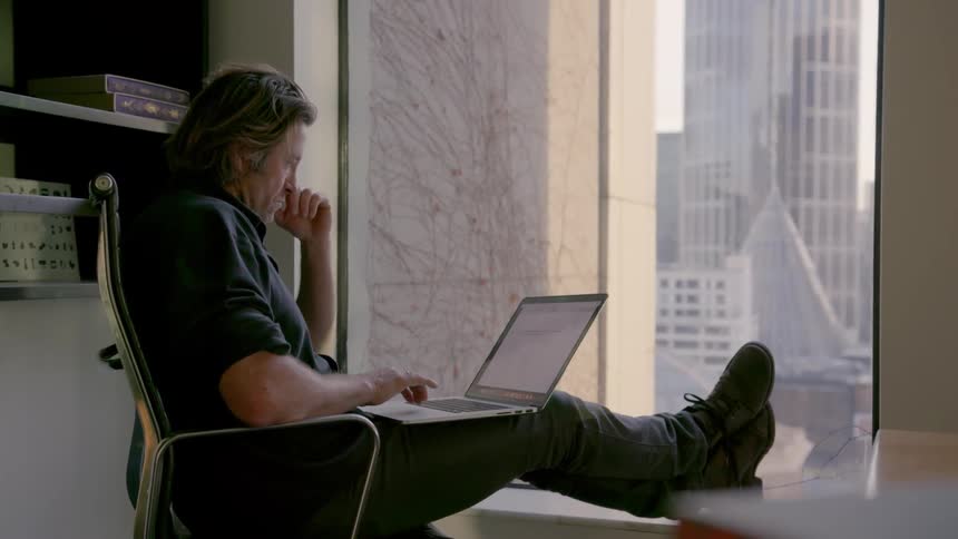 A man works on his laptop while sitting looking out a window in an office building