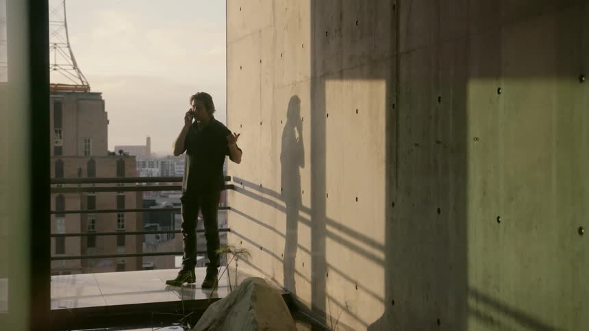 A man is standing on the ledge of a balcony and is talking to someone on the phone