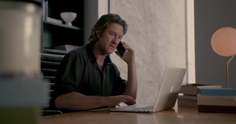A man is on the phone using his laptop at his desk