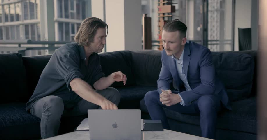 Two men are sitting on a couch and one man is showing the other something on the laptop