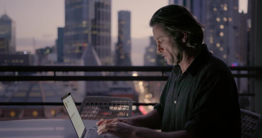 Man sitting at balcony table with city view is typing on laptop