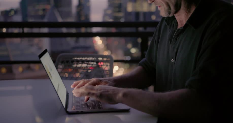 A man types on a laptop on a city balcony