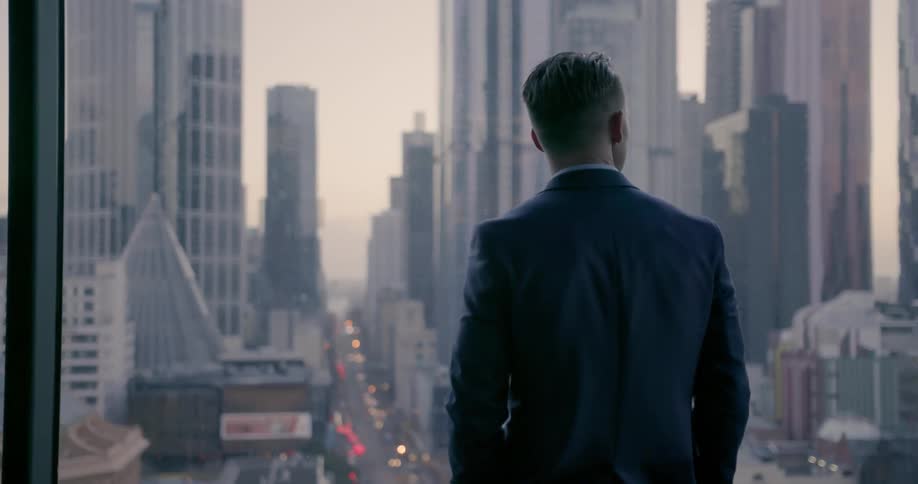 A man looks out of his large building window overlooking the busy city