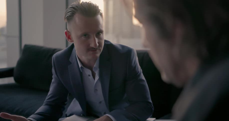 A man sits down with a book in his lap discussing something with another man