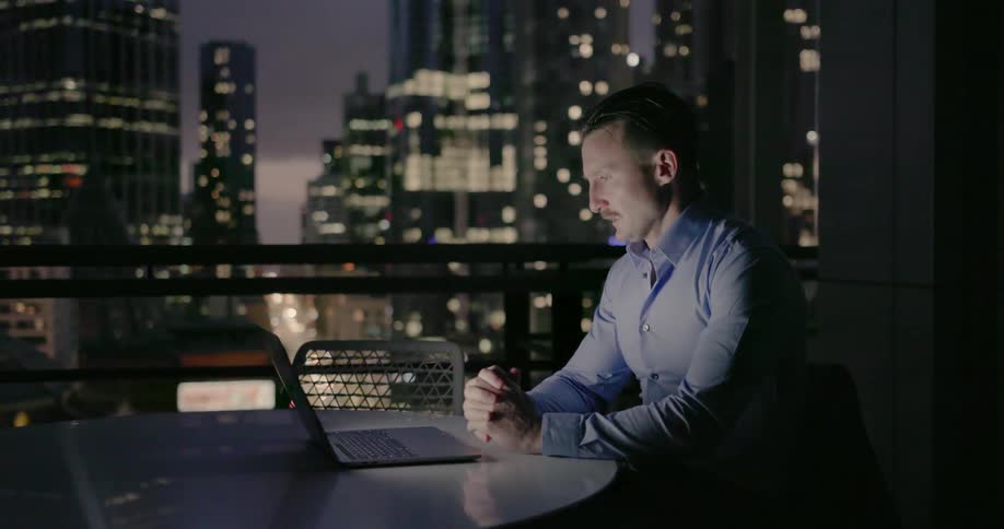 A man is sitting next to a window overlooking the city and he's talking to someone on his laptop