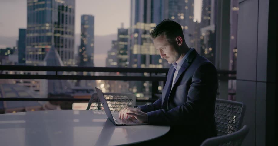 A businessman is working at a laptop while sitting on a balcony at dusk