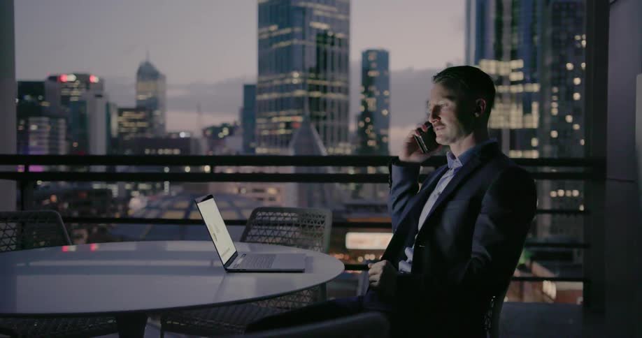 A businessman is working using a laptop and cellphone while sitting on a balcony at dusk