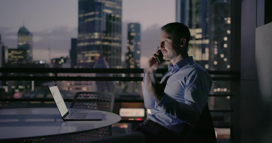 A man is talking on his phone and using his laptop with an open window view of a big city
