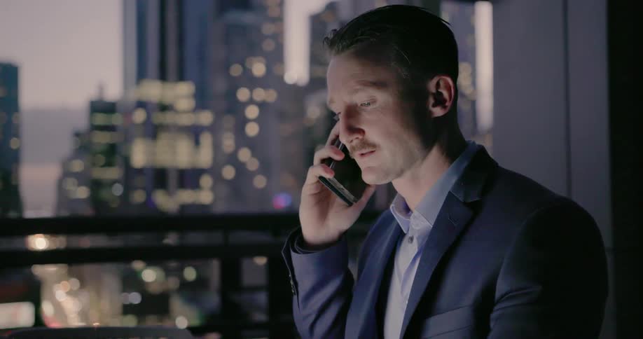 A businessman is working using a laptop and cellphone while sitting on a balcony at dusk