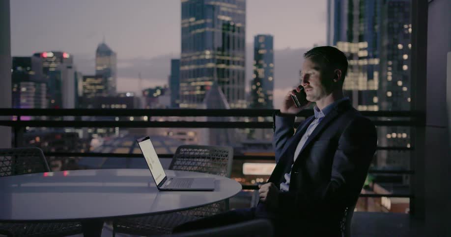 A businessman sits at a table on a balcony talking on his cellphone at dusk