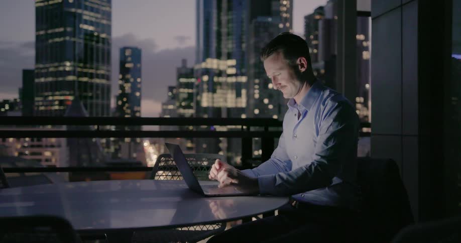 A businessman is working at a laptop while sitting on a balcony at dusk
