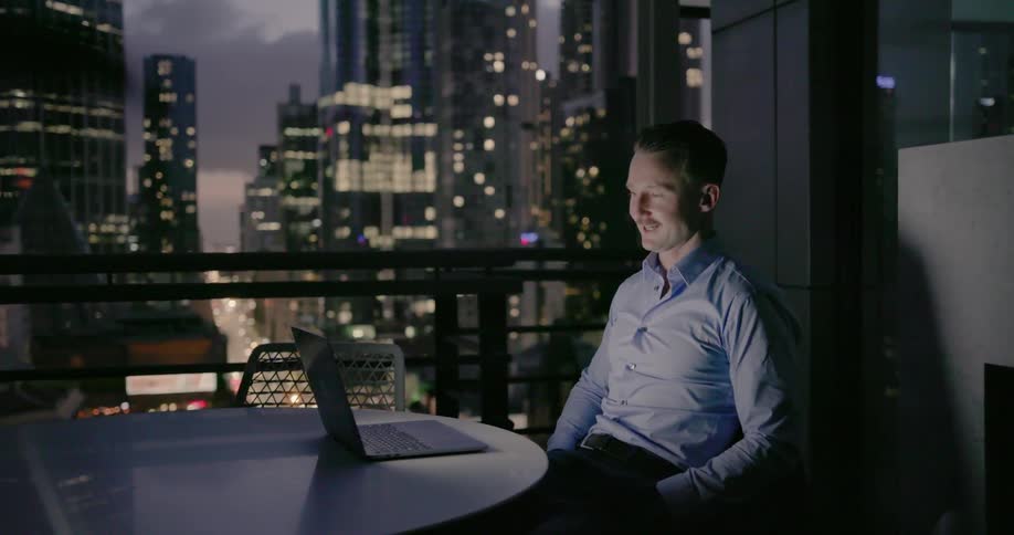 A man is sitting in his apartment talking to someone on the laptop and waving, the window is open and you can see the city in the background