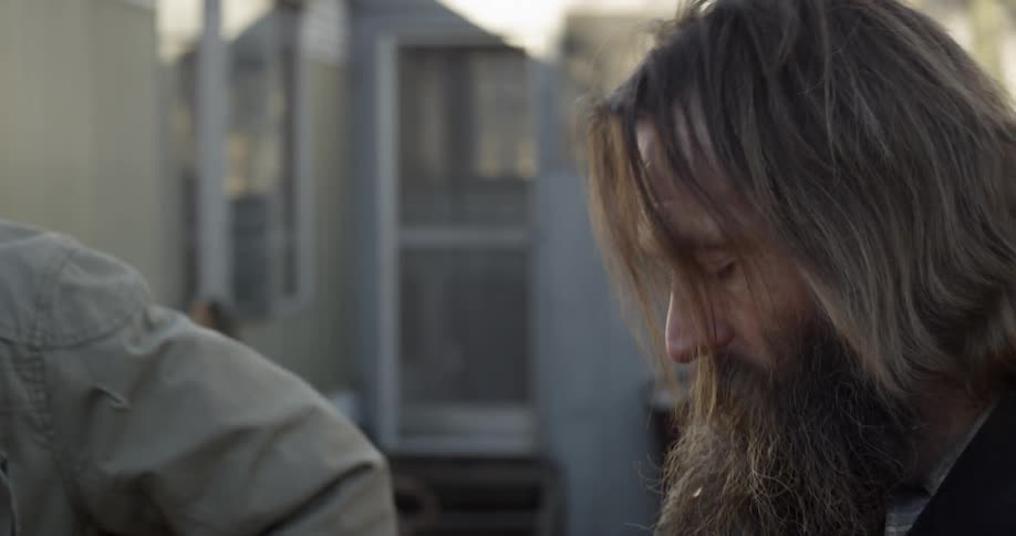 A young man approaches a bearded man outside a trailer
