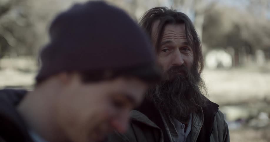 A man with a long beard is sitting and talking to a younger man wearing a hoodie