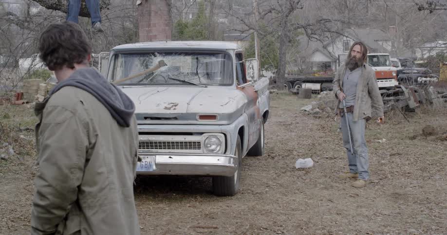 An older man carrying a rifle throws something to a teenager as they stand in a rural yard