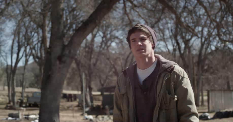 A young man standing in a rural yard speaks and then angrily walks away