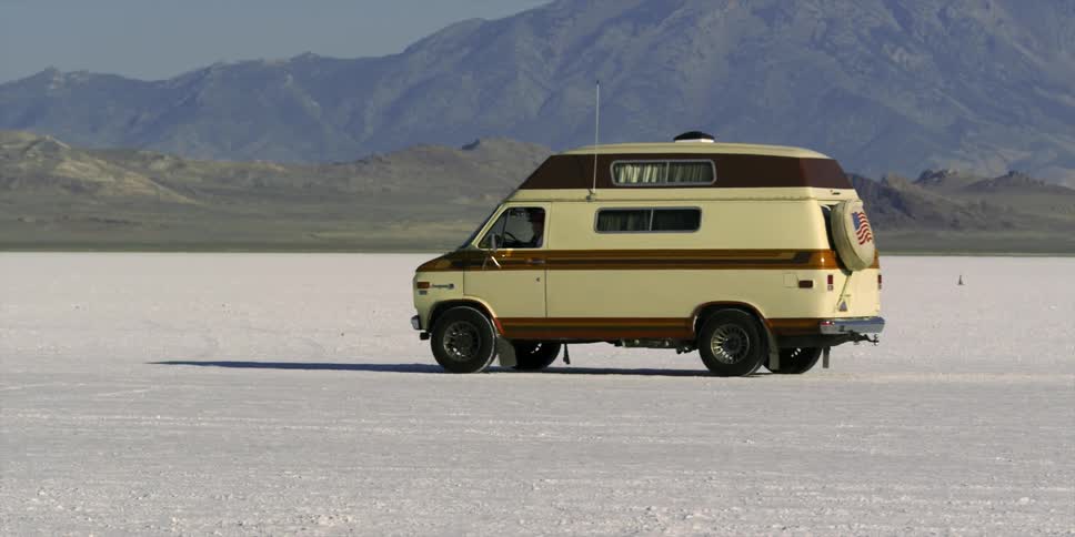 An old van conversion is driving across a salt flat in the desert during the day