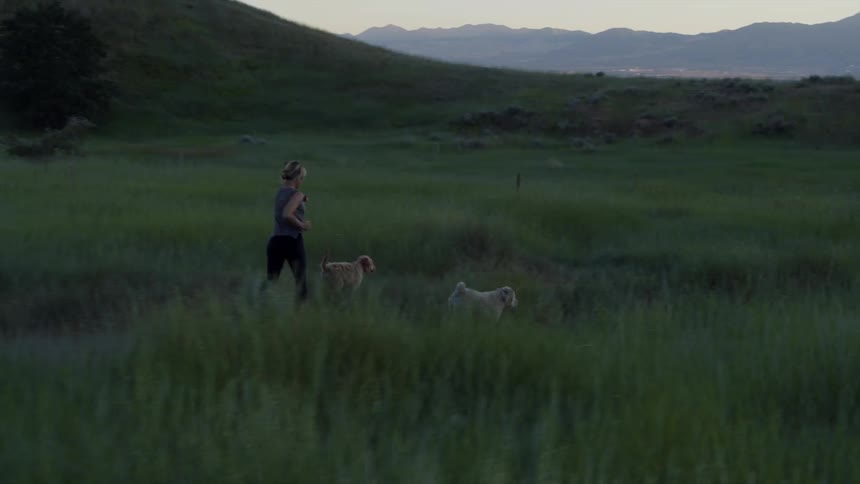 A woman running by some large hills through tall grass with two dogs at dusk