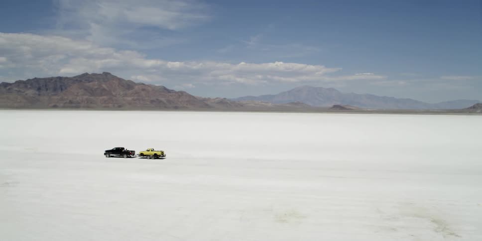Black truck pulls a yellow truck across a flat area of land