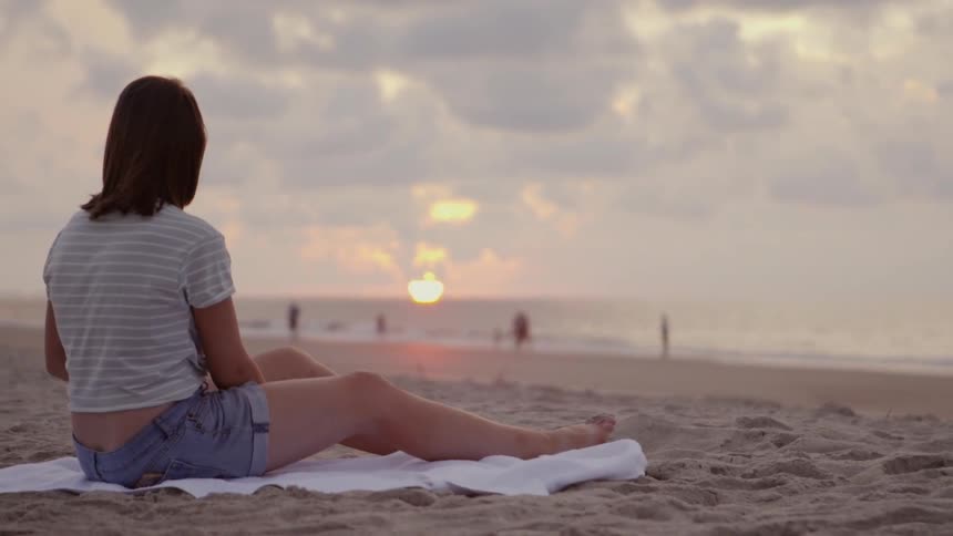 A girl is sitting on a towel watching the sunset at the beach