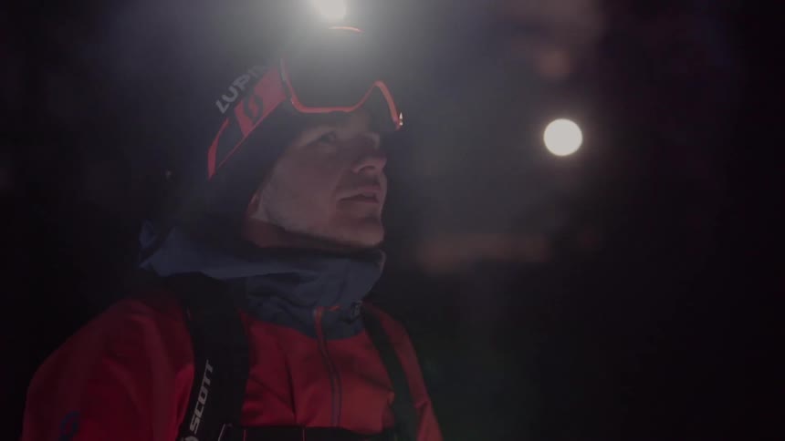 A man wearing a snow jacket and goggles with a headlamp removes them and looks around