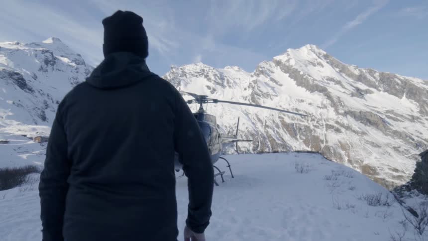 A man is walking down a snowy path towards a helicopter with mountains in the background