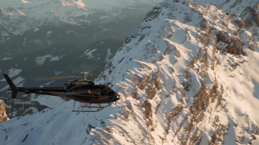 A helicopter flies over snowy mountains at sunset